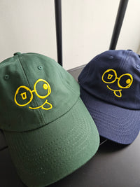 Green and navy dad hats displayed side by side, showing yellow glasses-face logos and stitched curved brims