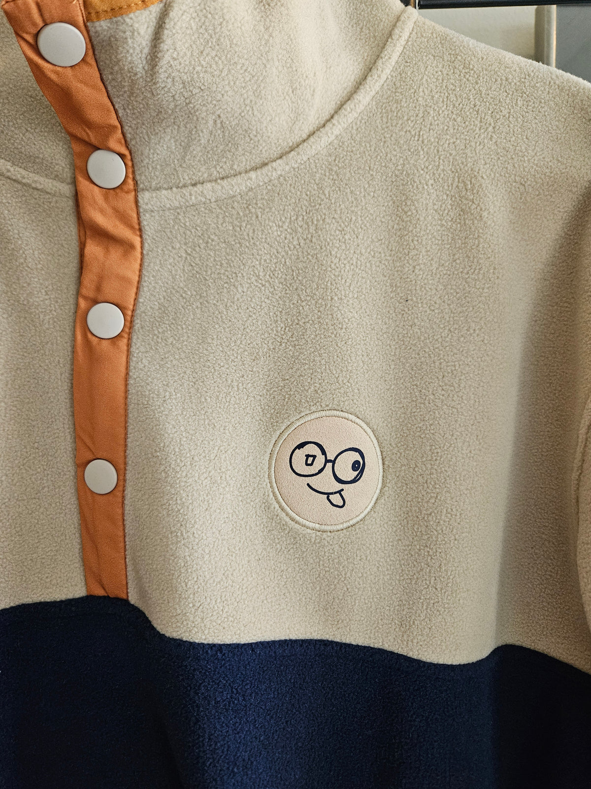 Close-up of beige fleece stand collar, orange placket with four snaps and round glasses-face chest patch