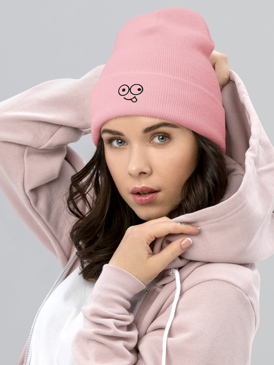 Model wearing a baby pink cuffed knit beanie with a black tongue-out glasses-face design