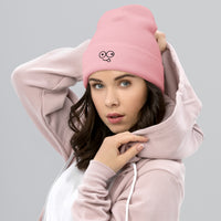 Model wearing a baby pink cuffed knit beanie with a black tongue-out glasses-face design