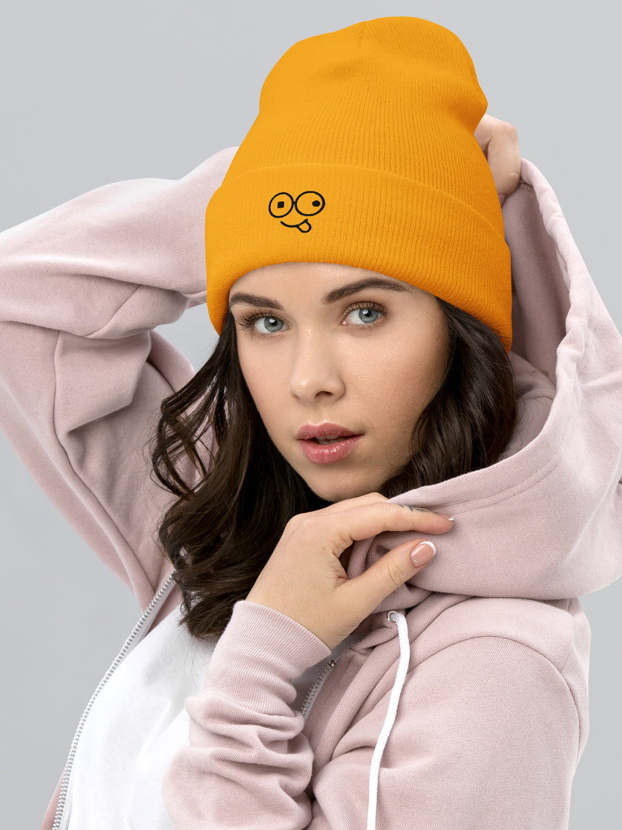 Model wearing a gold cuffed knit beanie with a black tongue-out glasses-face design