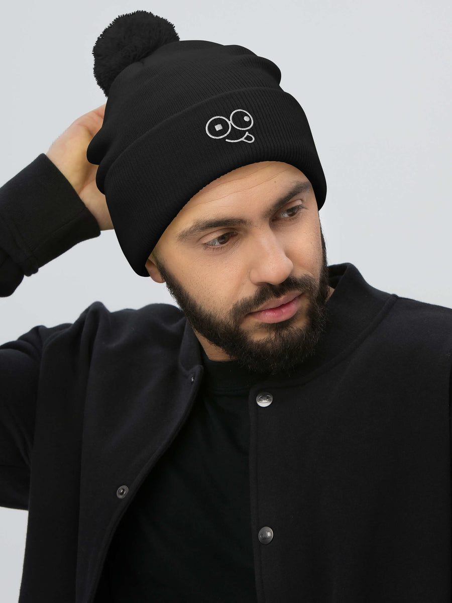 Model wearing a black pom-pom knit beanie with a folded cuff and white tongue-out glasses-face design