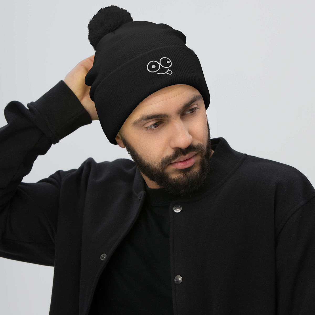 Model wearing a black pom-pom knit beanie with a folded cuff and white tongue-out glasses-face design