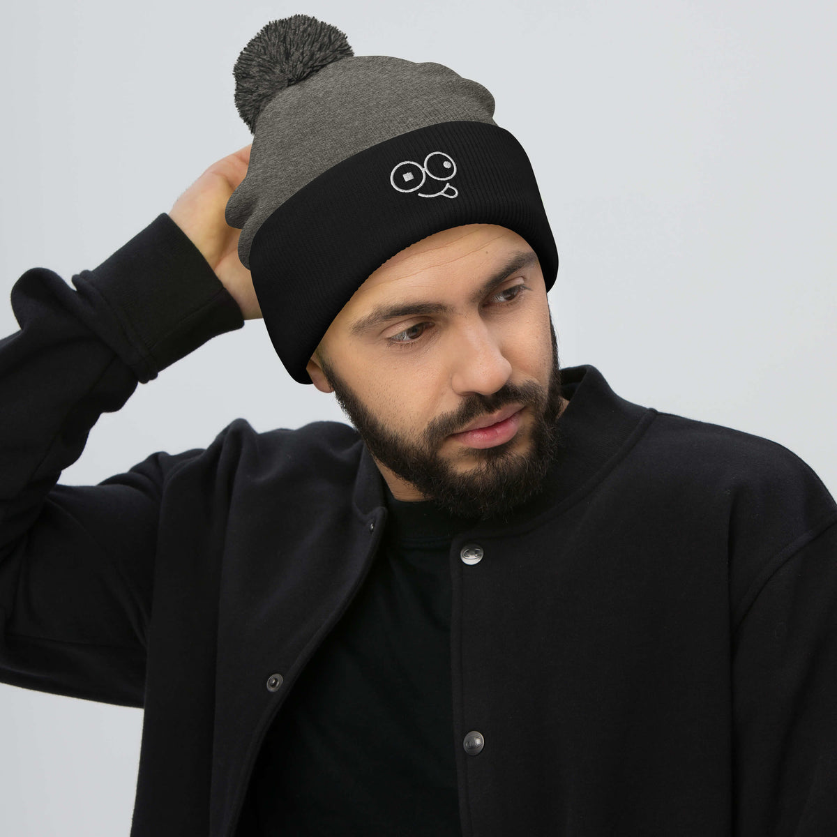 Model wearing a dark heather grey pom-pom beanie with a black cuff and white tongue-out glasses-face design