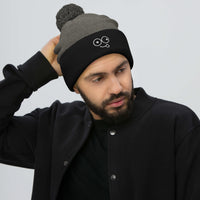 Model wearing a dark heather grey pom-pom beanie with a black cuff and white tongue-out glasses-face design