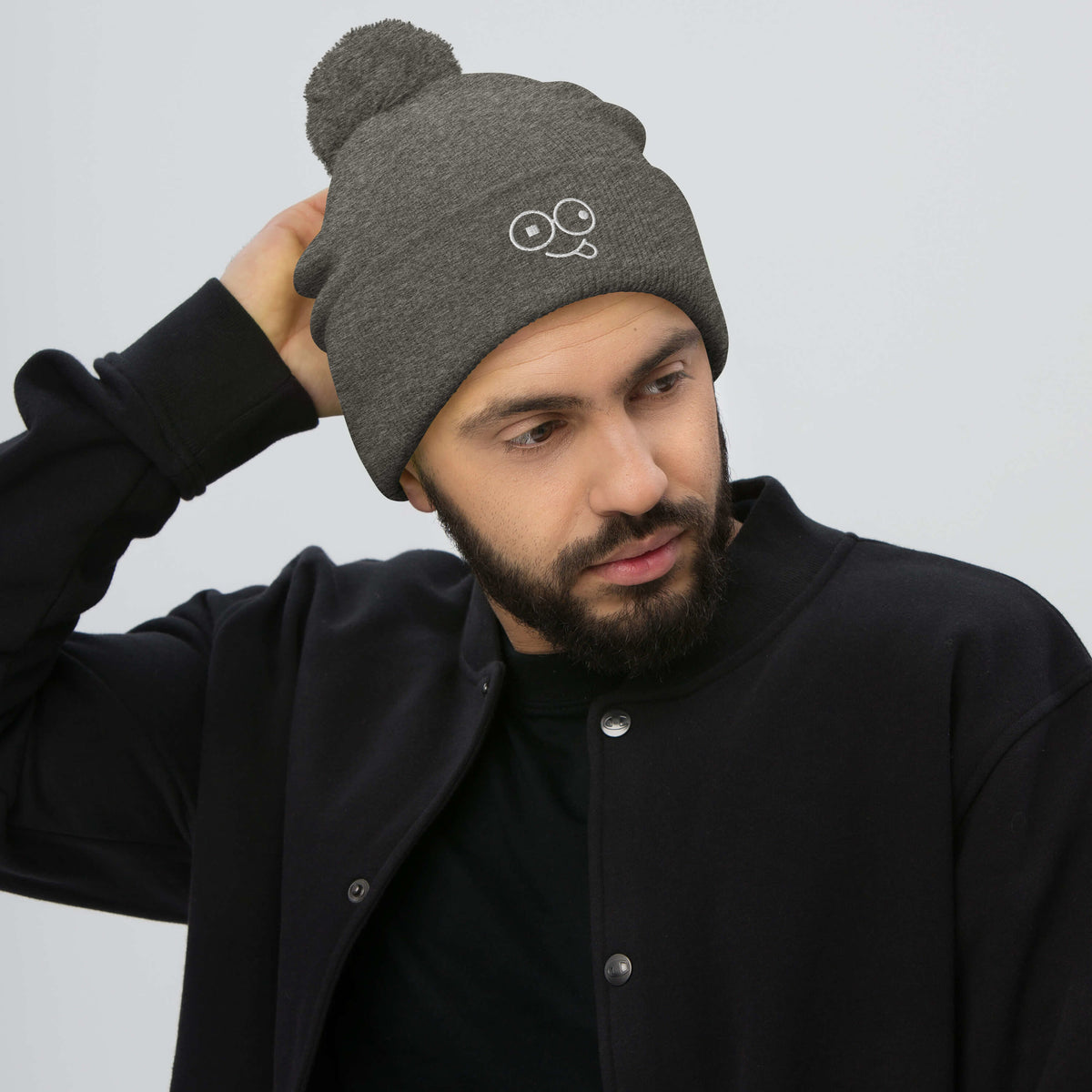 Model wearing a dark heather grey pom-pom knit beanie with a folded cuff and white tongue-out glasses-face design