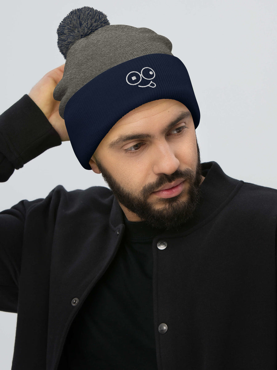 Model wearing a dark heather grey pom-pom beanie with a navy cuff and white tongue-out glasses-face design