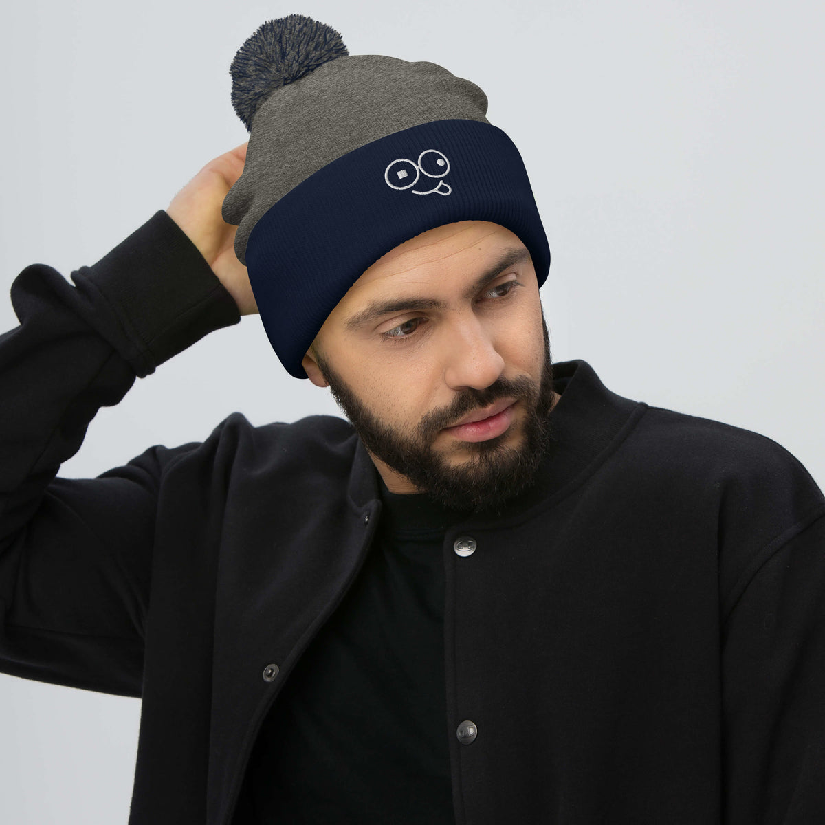 Model wearing a dark heather grey pom-pom beanie with a navy cuff and white tongue-out glasses-face design