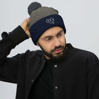 Model wearing a dark heather grey pom-pom beanie with a navy cuff and white tongue-out glasses-face design