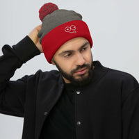 Model wearing a dark heather grey beanie with a red cuff, two-tone pom-pom and tongue-out glasses-face design