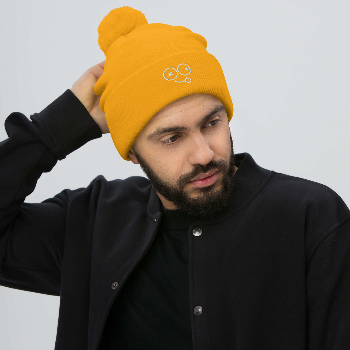 Model wearing a gold pom-pom knit beanie with a folded cuff and light-colored tongue-out glasses-face design