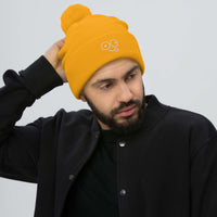 Model wearing a gold pom-pom knit beanie with a folded cuff and light-colored tongue-out glasses-face design