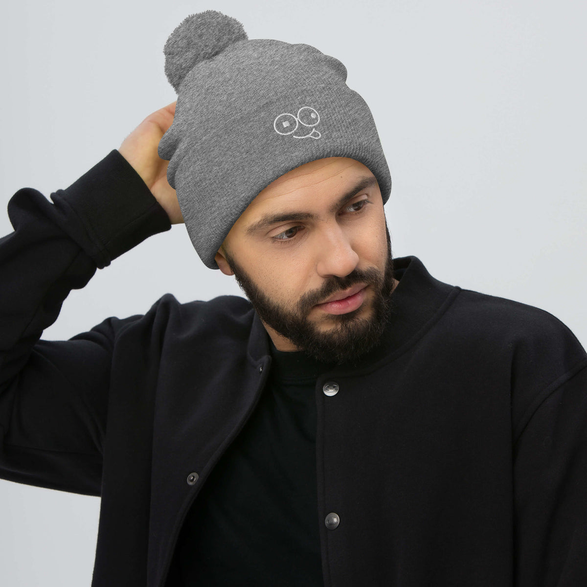 Model wearing a light heather grey pom-pom knit beanie with a folded cuff and white tongue-out glasses-face design