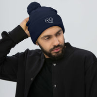 Model wearing a navy pom-pom knit beanie with a folded cuff and white tongue-out glasses-face design