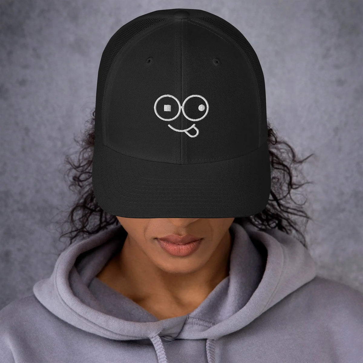 Model wearing a black trucker hat with a curved brim and white tongue-out glasses-face design