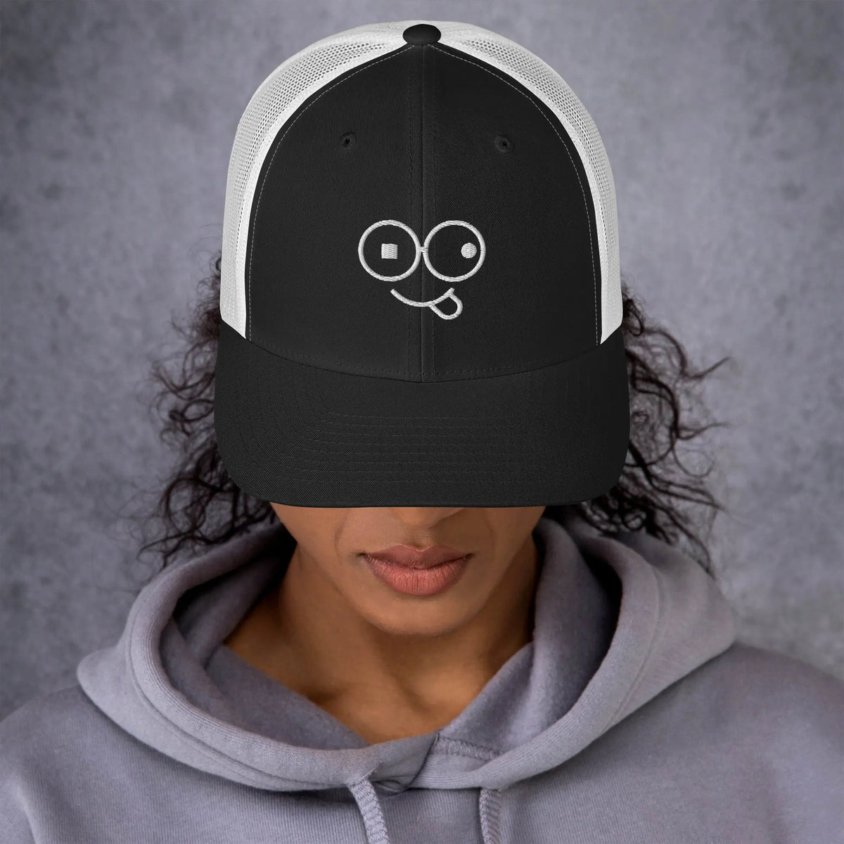 Model wearing a black trucker hat with white mesh panels and a white tongue-out glasses-face design