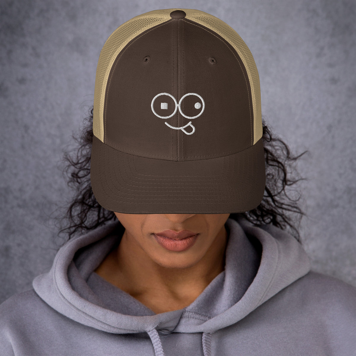 Model wearing a brown trucker hat with khaki mesh panels and a white tongue-out glasses-face design