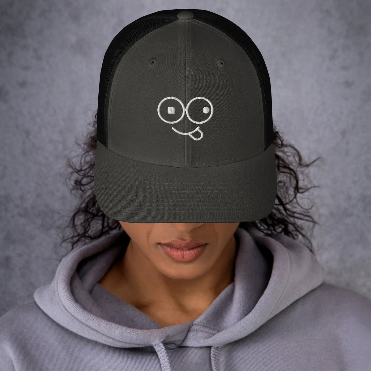 Model wearing a charcoal trucker hat with black mesh panels and a white tongue-out glasses-face design