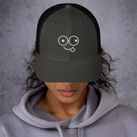 Model wearing a charcoal trucker hat with black mesh panels and a white tongue-out glasses-face design