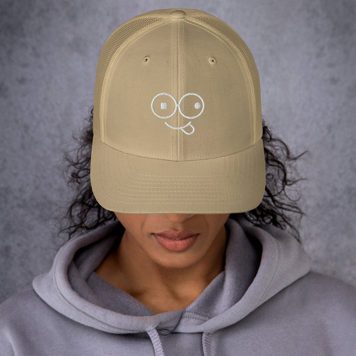 Model wearing a khaki trucker hat with a curved brim and white tongue-out glasses-face design