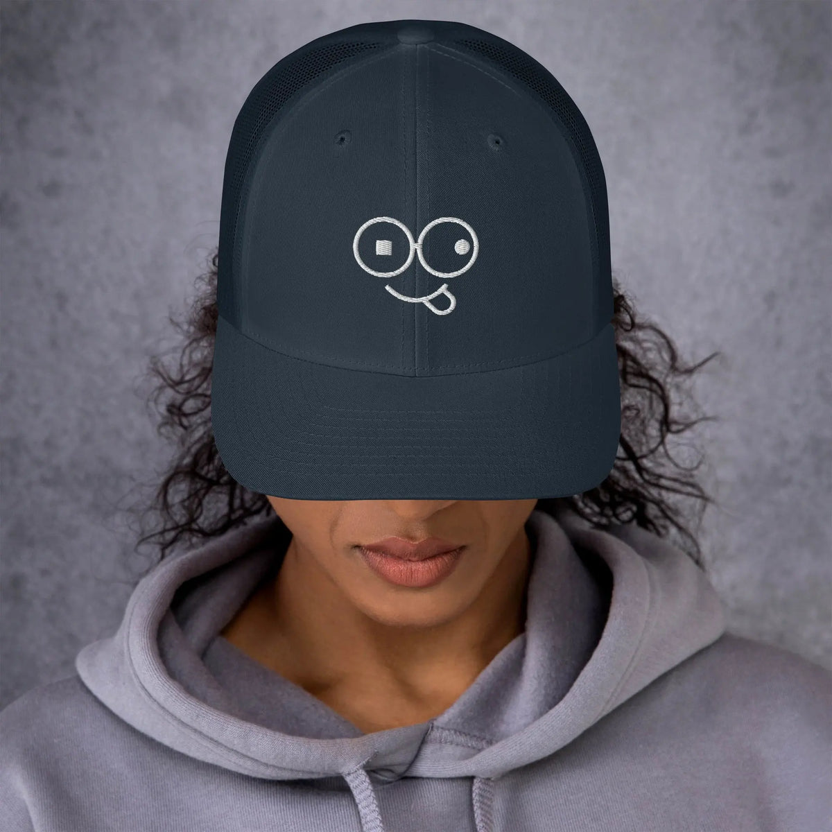 Model wearing a navy trucker hat with a curved brim and white tongue-out glasses-face design