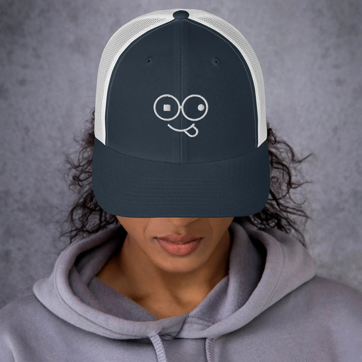 Model wearing a navy trucker hat with white mesh panels and a white tongue-out glasses-face design