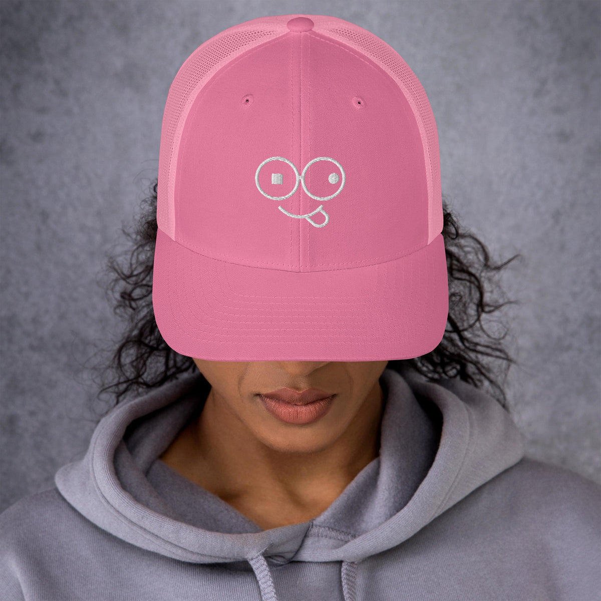 Model wearing a pink trucker hat with a curved brim and white tongue-out glasses-face design