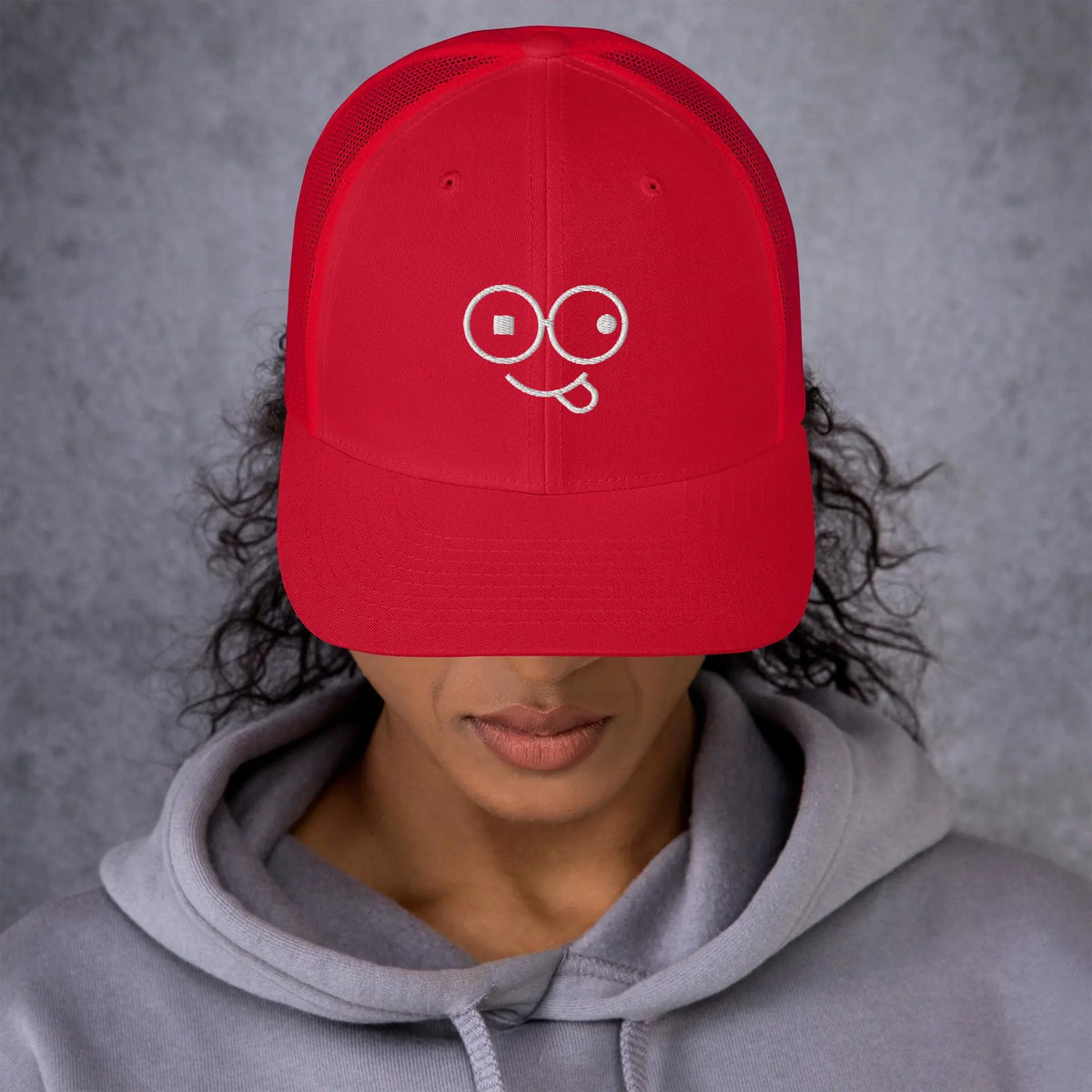 Model wearing a red trucker hat with a curved brim and white tongue-out glasses-face design