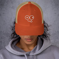 Model wearing a rustic orange trucker hat with khaki mesh panels and a white tongue-out glasses-face design