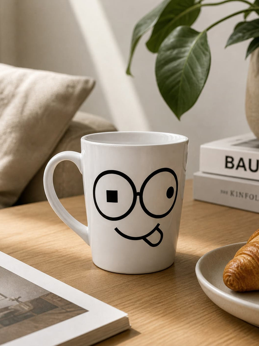 White funny face coffee mug with black glasses and a tongue-out smile on a wooden table beside a croissant