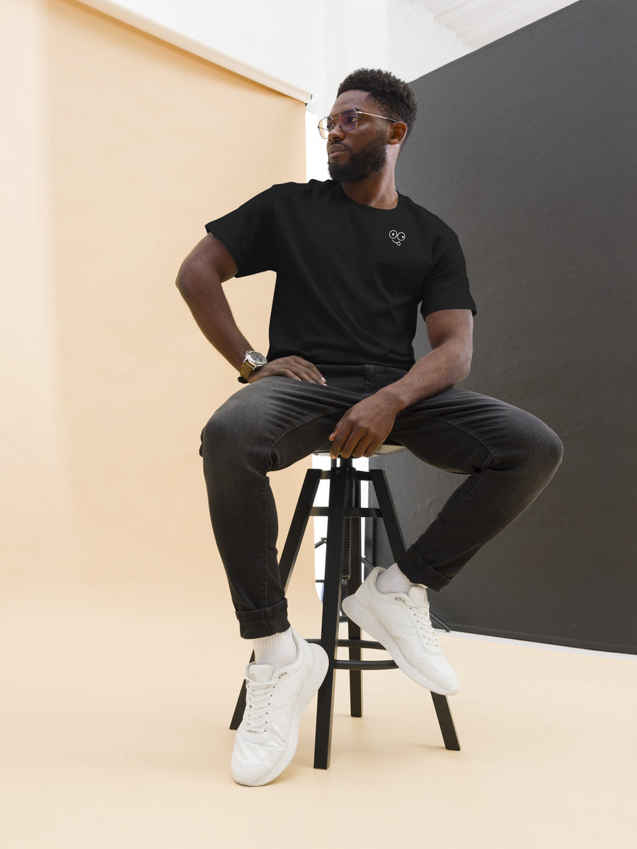 Model seated on a stool wearing a black short sleeve t-shirt with a white glasses-face chest design