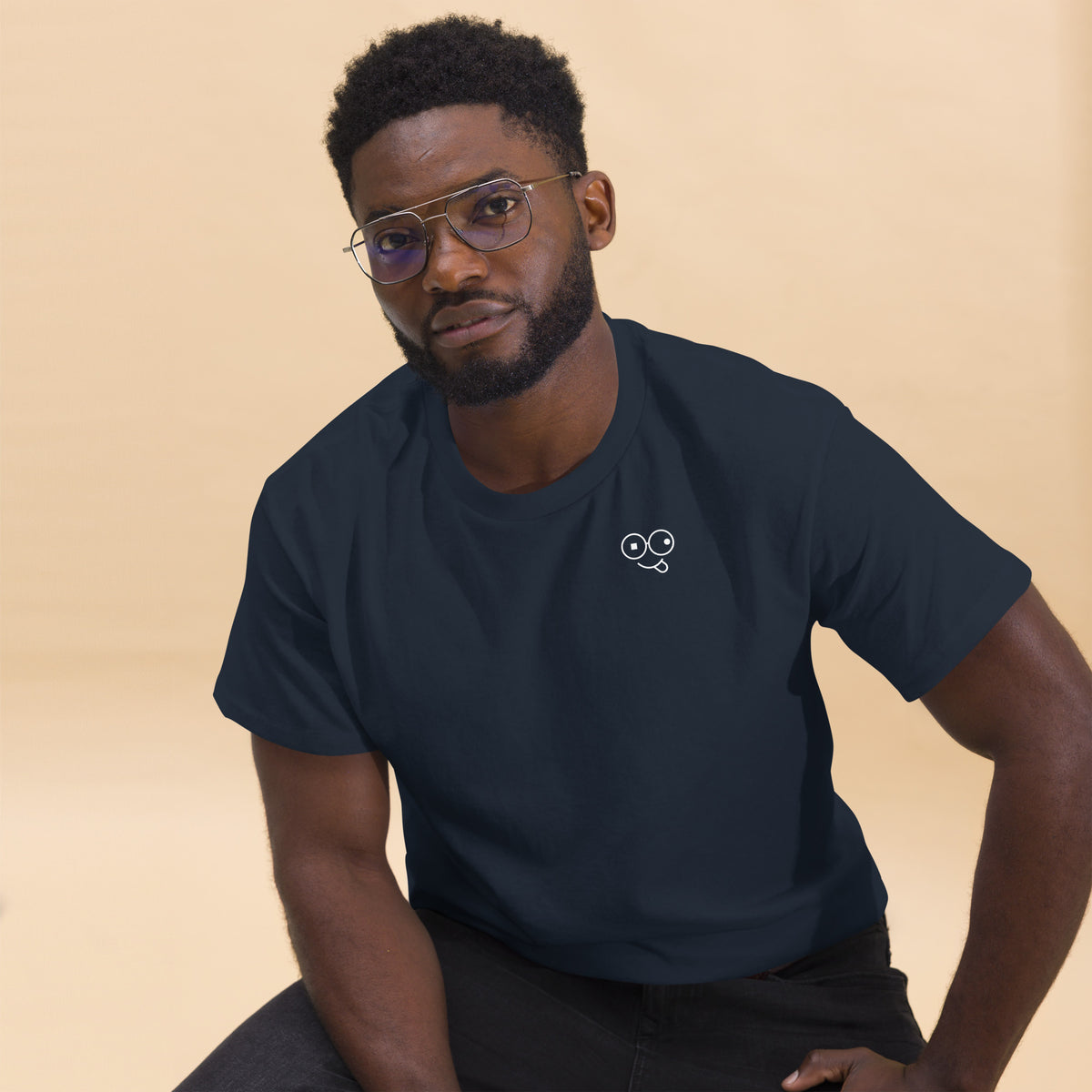 Seated model wearing a navy crew neck t-shirt with a small white tongue-out glasses-face chest design