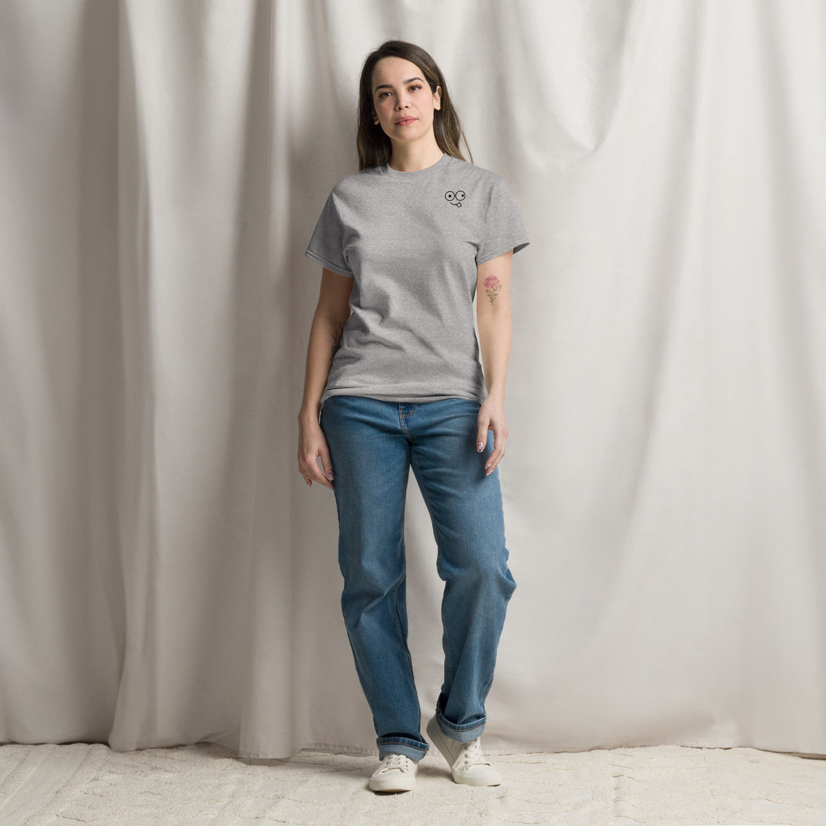 Model wearing a sport grey crew neck t-shirt with a small black glasses-face chest design and blue jeans