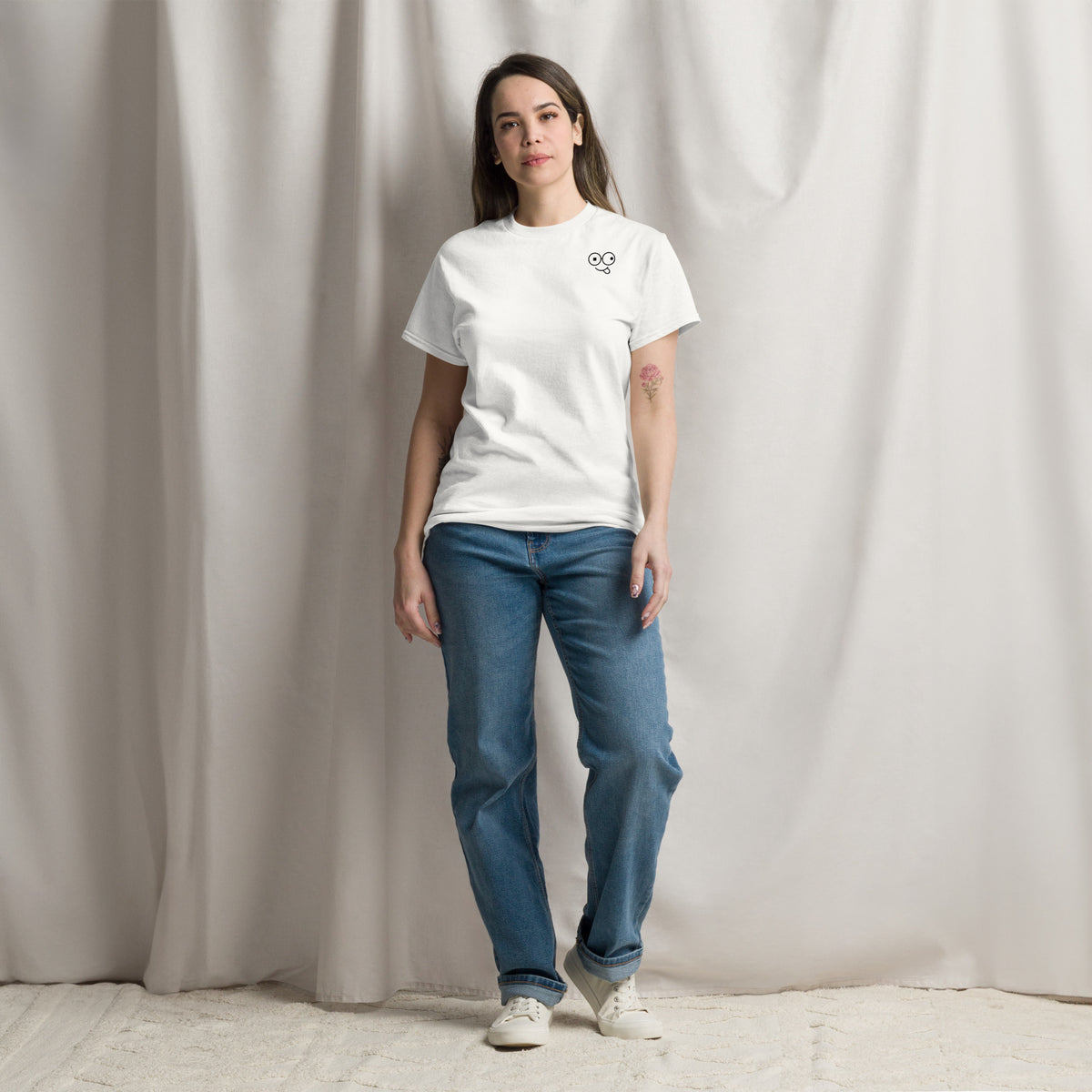 Model wearing a white crew neck t-shirt with a small black glasses-face chest design and blue jeans