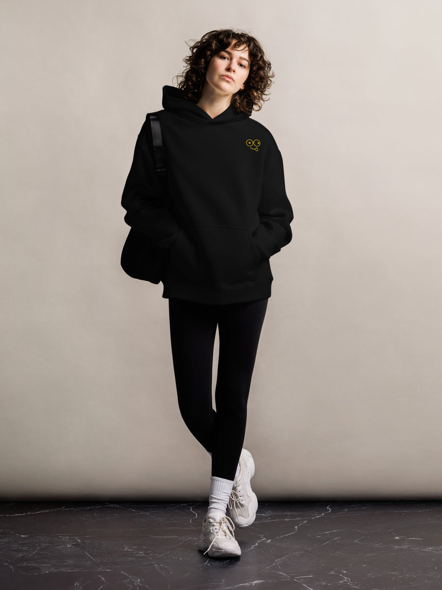 Model wearing a black oversized pullover hoodie with a small yellow glasses-face chest design and front kangaroo pocket