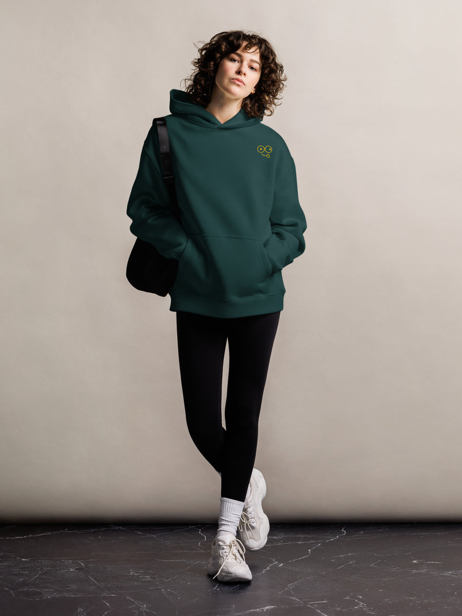 Model wearing a black oversized pullover hoodie with a small yellow glasses-face chest design and front kangaroo pocket