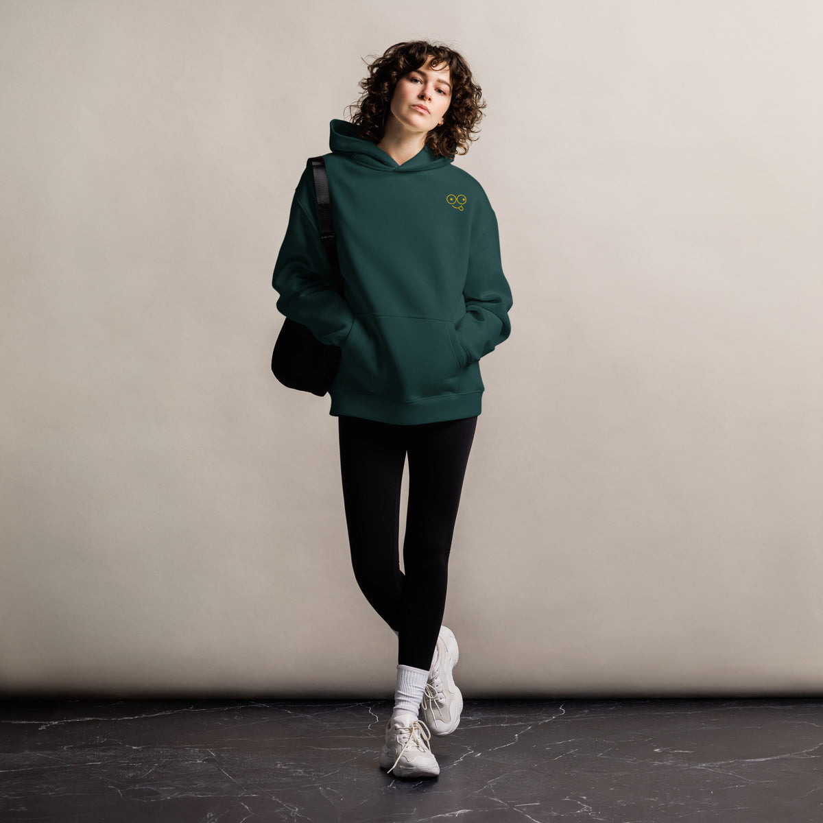 Model wearing a black oversized pullover hoodie with a small yellow glasses-face chest design and front kangaroo pocket