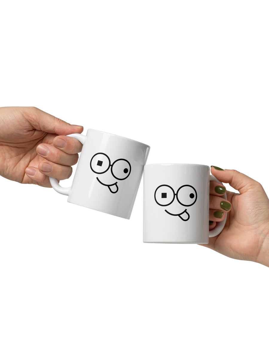 Two white mugs held together in a toast, each featuring black round glasses, mismatched eyes and a tongue-out smile