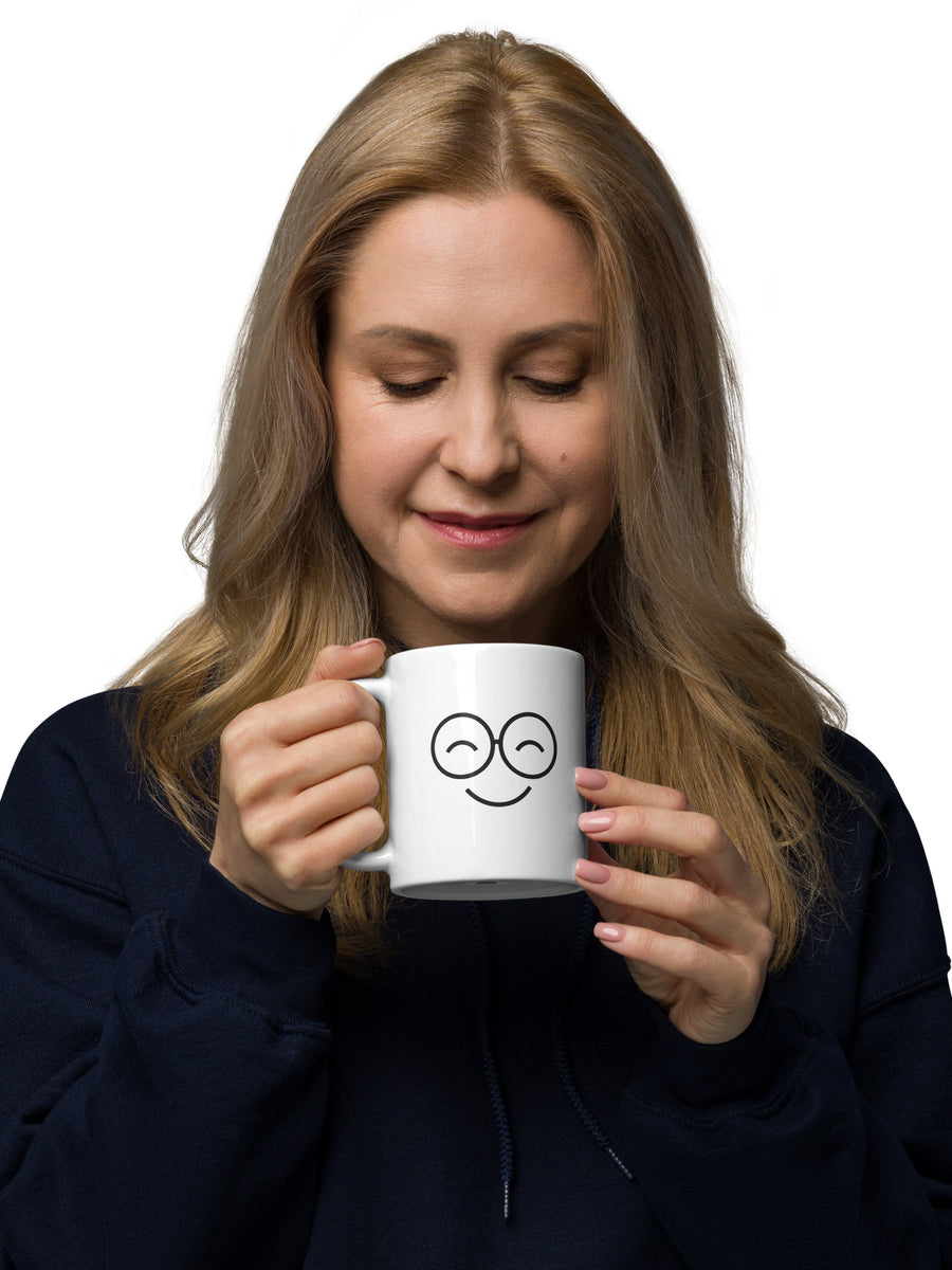 Person holding a white mug with a smiling glasses-face design and closed eyes, with the handle on the left