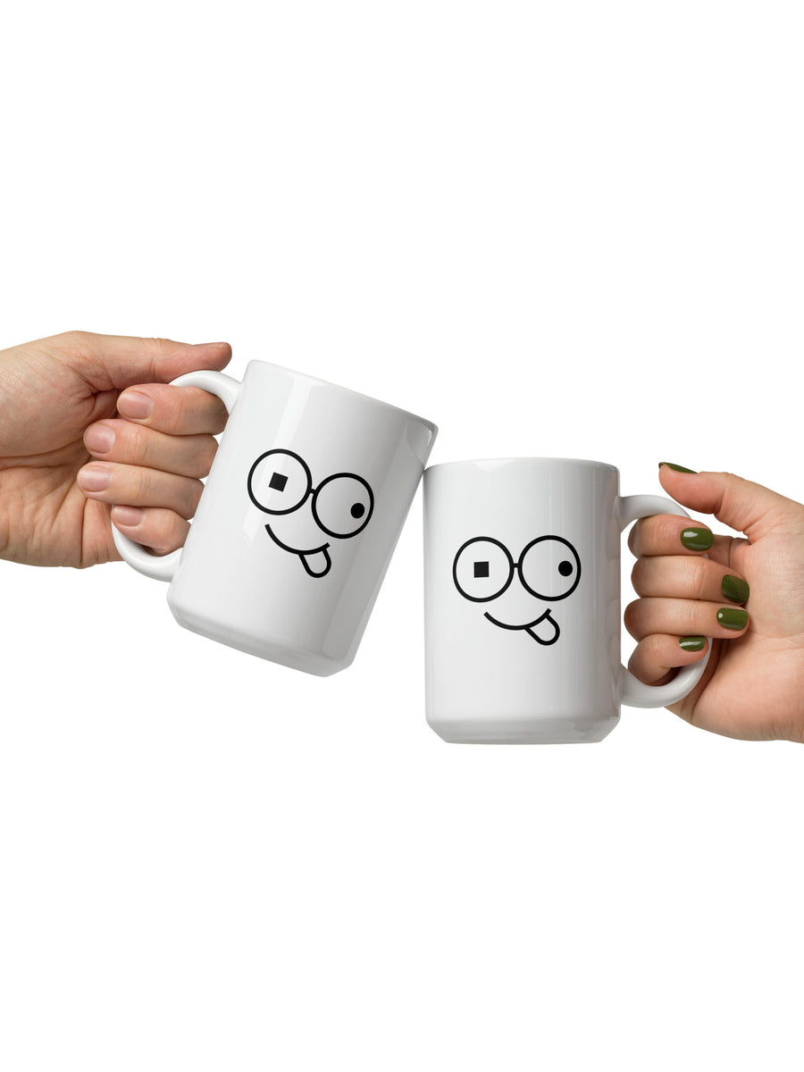 Pair of white mugs with black glasses-face artwork and tongue-out smiles, held by their handles against a white background